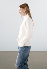 White button-up shirt with a high collar, long sleeves, and straight fit, paired with light blue jeans. Smooth fabric, simple design.