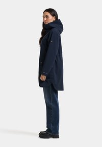 Femme debout de profil portant une longue veste à capuche bleu marine foncé, un jean bleu et des bottes noires sur un fond clair uni.