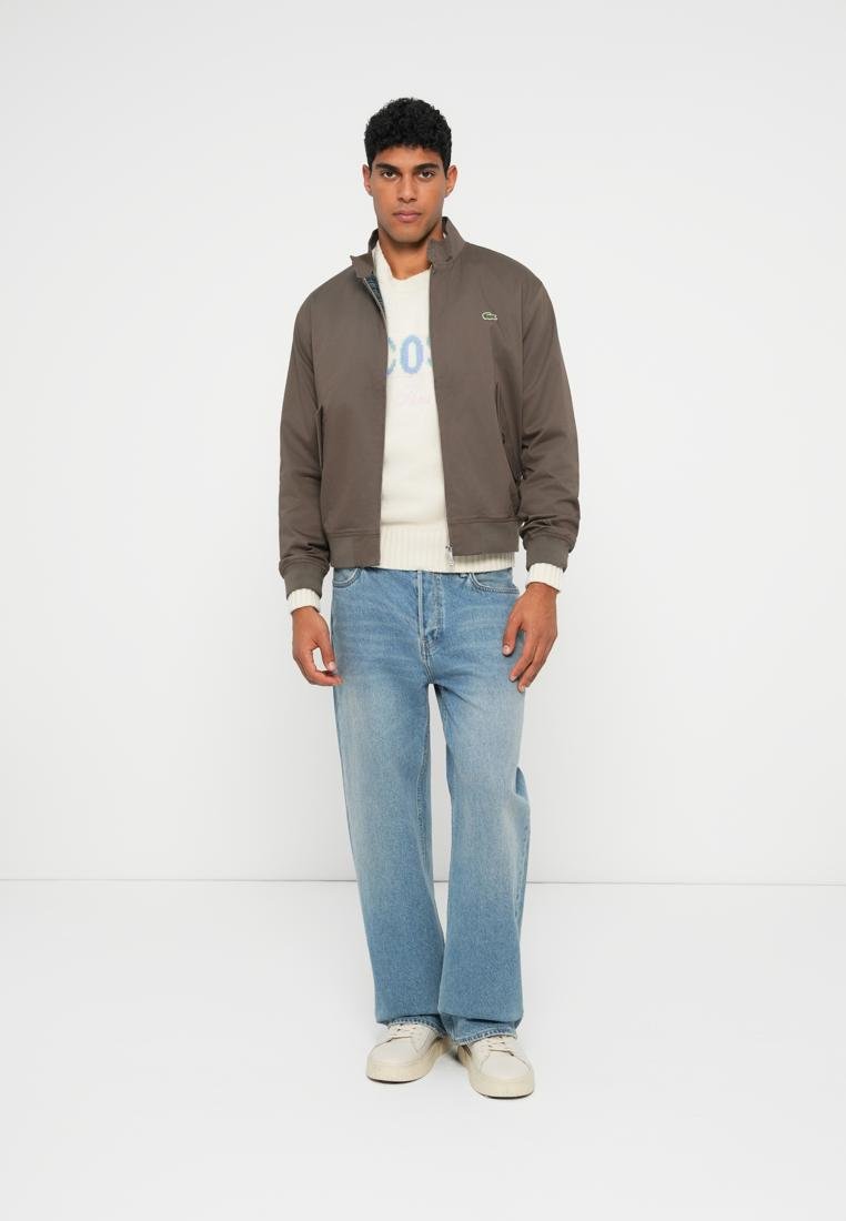 Veste aviateur marron avec fermeture éclair, associée à un pull crème et un jean large bleu clair. Des baskets blanches complètent la tenue.