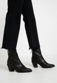 Botas de tobillo de cuero negro con textura de cocodrilo, punta afilada y un tacón grueso. Combinadas con jeans negros cortos con dobladillos deshilachados.
