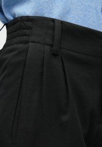 Prim-plan al pantalonilor negri cu talie înaltă, având pliuri și bucle pentru curea, purtați cu un top tricotat de culoare albastru deschis.
