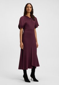 Donna con lunghi capelli scuri che indossa un vestito midi prugna con maniche a sbuffo e stivali neri con tacco, in piedi davanti a uno sfondo bianco semplice.