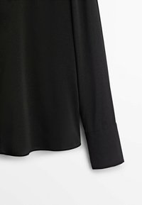 Chemise noire à manches longues avec larges poignets et ourlet droit, montrée de la taille au poignet sur un fond blanc.