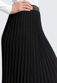 Jupe plissée noire avec une taille texturée. Le tissu est lisse et les plis créent une silhouette fluide. Accentuée d'une bande brillante.