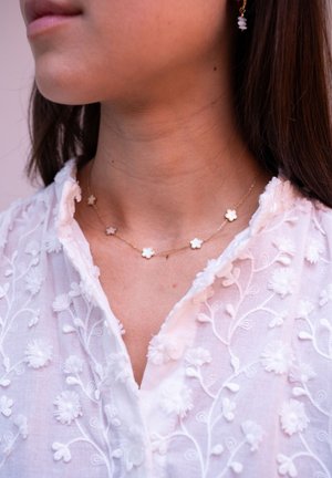 Złoty naszyjnik z łańcuchem z białymi pendantami w kształcie kwiatów. Posiada pięć płatków, delikatny design, noszony z białą haftowaną bluzką.