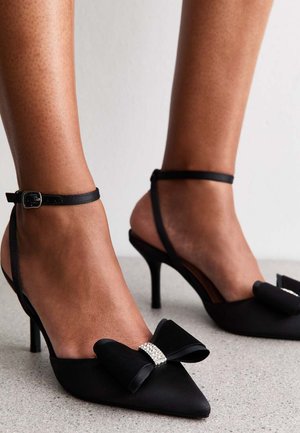 Zwarte satijnen schoenen met hoge hak en puntige neus, voorzien van een grote strik en steendetail aan de voorkant, en enkelbandjes.