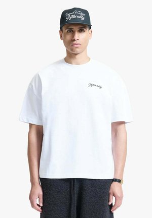 Uomo che indossa un cappellino nero con testo bianco, una maglietta bianca oversize con piccolo testo nero sul petto e pantaloni scuri con trama, su sfondo bianco.