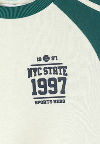 Valge ja roheline raglaan-särk tumesinise tekstiga "NYC State 1997 Sports Hero" ning väikese tennispalli graafikuga numbrite 19 ja 97 vahel.