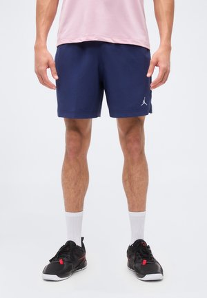 Uomo che indossa pantaloncini sportivi Jordan blu navy, maglietta rosa, calzini bianchi e sneakers nere, in piedi su sfondo bianco.