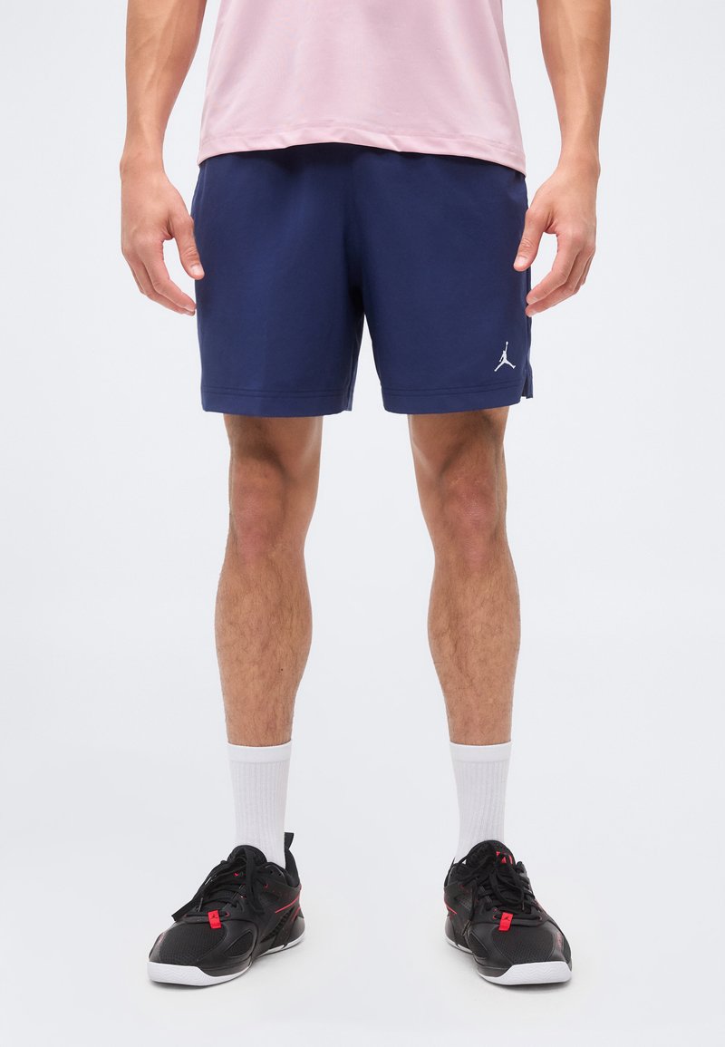 Homme portant un short de sport Jordan bleu marine, un t-shirt rose, des chaussettes blanches et des baskets noires, debout sur fond blanc.