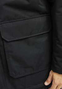 Chaqueta negra con un gran bolsillo rectangular, tejido texturizado y acentos cosidos. El primer plano muestra una mano agarrando el dobladillo cerca del bolsillo.