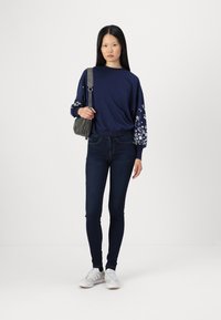 Marinblå sweatshirt med blommönster på ärmarna, ihopparad med mörka skinny jeans och vita sneakers. Modellen håller en grå handväska.