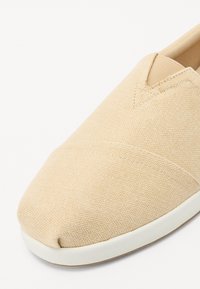 Chaussure slip-on beige avec un motif chevron texturé, un bout rond, des inserts élastiques sur les côtés et une semelle en caoutchouc blanc contrastant.