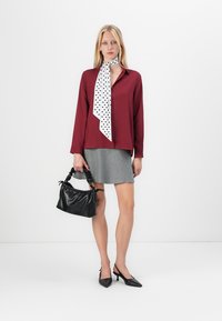 Burgundy skjorta med lång ärm och knappar, grå kjol, prickig scarf och svart texturerad handväska. Spetsiga svarta klackar fulländar looken.