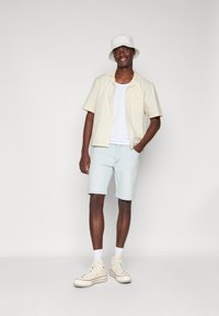Camisa de manga corta color crema, top blanco de tirantes, pantalones cortos de mezclilla azul claro, zapatillas altas color crema y un sombrero tipo bucket blanco.