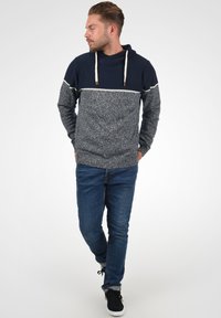 Haut en tricot à capuche bleu marine et gris avec une section inférieure texturée, une bande horizontale blanche et des cordons de serrage. Associé à un jean bleu et des baskets noires.