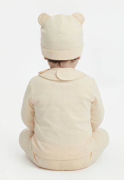 Bebé con un conjunto beige suave y un gorro a juego con orejas de oso, sentado con las piernas cruzadas de espaldas sobre un fondo blanco.