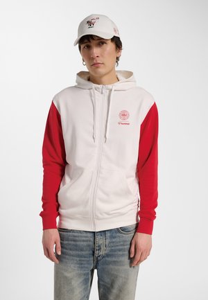 Ung mand iført en hvid og rød hættetrøje med lynlås med DBU-logo og lyseblå jeans, stående mod en ensfarvet hvid baggrund.