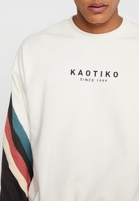 Man som bär en vit sweatshirt med texten "KAOTIKO SINCE 1999" på bröstet och flerfärgade ränder på vänster ärm.