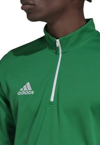 Grünes Sportoberteil mit halbem Reißverschluss aus strukturiertem Stoff, mit einem weißen Reißverschluss und einem weißen gestickten Logo auf der linken Brust.