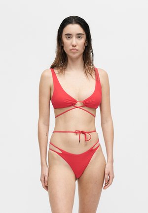 Γυναίκα με βρεγμένα μαλλιά που φοράει κόκκινο μπικίνι με λεπτομέρειες τύλιγμα και δεσίματος, στέκεται μπροστά σε απλό λευκό φόντο.