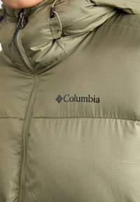 Grön Columbia-pufferjacka med quiltad textur, dragkedja och logotyp. Materialet har ett subtilt rutmönster och en hög krage.