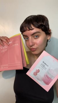 Twee roze en gele verpakkingen van huidverzorgingsmaskers worden omhoog gehouden door een persoon, met een gladde, glanzende verpakking met minimale tekst en grafische accenten.