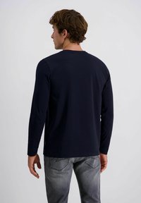 Mann mit braunen Haaren, der ein dunkelblaues Langarmshirt und ausgewaschene graue Jeans trägt, steht mit dem Rücken zu einem schlichten hellen Hintergrund.