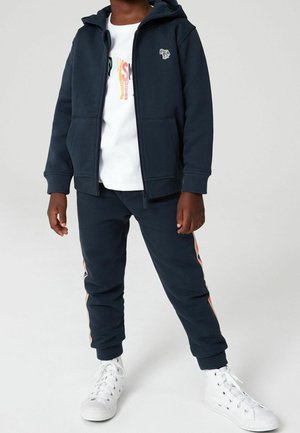 Granatowa kurtka z kapturem i przednimi kieszeniami, w zestawie z dopasowanymi joggerami z pomarańczowymi bocznymi paskami. Całość dopełniają białe sneakersy.