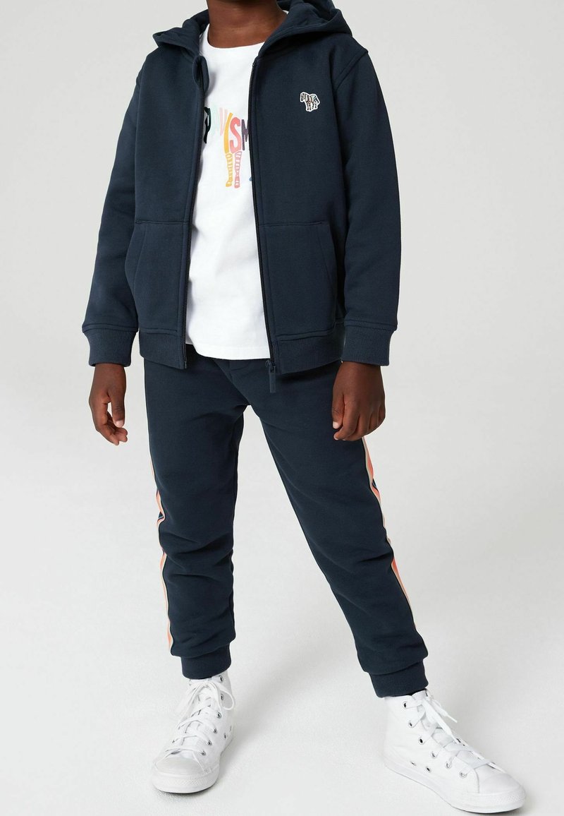 Veste à capuche bleu marine avec poches avant, associée à un jogging assorti avec des bandes latérales orange. Des baskets blanches complètent la tenue.