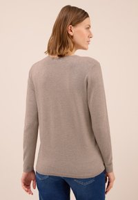 Žena s vlasy po ramena, oblečená v béžovém svetr s dlouhým rukávem a modrých džínách, stojící zády k jednoduchému světlému pozadí.