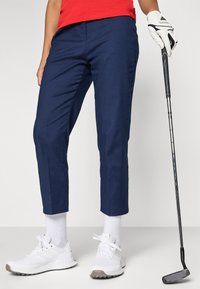 Persona che indossa pantaloni blu navy, sneakers bianche e una maglietta rossa, tiene in mano un bastone da golf con un guanto bianco su una mano.