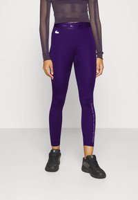 Leggings morados con un diseño ajustado, una textura suave y un eslogan impreso a lo largo del lado. Se llevan con una camiseta de manga larga de malla y zapatillas negras.