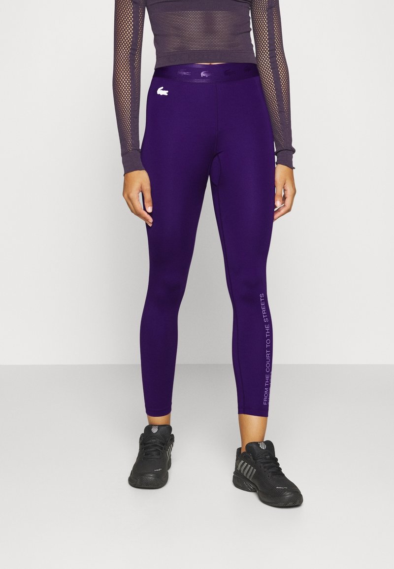 Leggings morados con un diseño ajustado, una textura suave y un eslogan impreso a lo largo del lado. Se llevan con una camiseta de manga larga de malla y zapatillas negras.