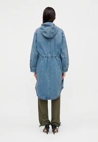 Μπουφάν από τζιν με κουκούλα, με σφηκοφωλιά στη μέση και μακριά μανίκια. Διαθέτει καμπύλη τελείωμα και εμφανή ραφές σε ανοιχτό μπλε χρώμα.