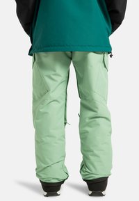Jasnozielone spodnie narciarskie o luźnym kroju, z kieszeniami cargo i zamkami na kostkach, noszone na czarnych butach.