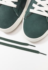 Baskets en daim vert avec des lacets blancs et une finition texturée. Deux lacets verts assortis sont placés sous les chaussures.