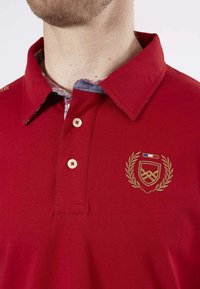 Polo rouge en coton. Présente un col, trois boutons et une broderie dorée avec un motif de bouclier et de lauriers. La doublure intérieure a un motif à carreaux.