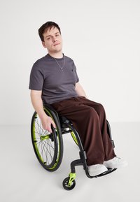 Μαύρο αναπηρικό καροτσάκι με φωτεινές πράσινες λεπτομέρειες. Το άτομο φοράει ένα γκρι T-shirt και καφέ παντελόνι. Είναι ορατά άσπρα παπούτσια. Απλό φόντο.