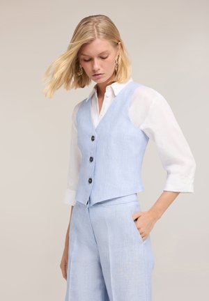 Kvinde iført lyseblå vest og matchende bukser med hvid skjorte, stående med den ene hånd i lommen, kigger ned.