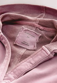 Ružičasta pamučna košulja s izlizanim etiketom koja glasi "THE EVERYDAY FAITHFUL NEXT, MADE IN BANGLADESH." Teksturirana tkanina s detaljima šivanja.