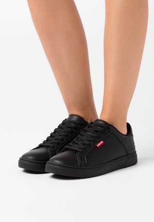 Zwarte lage sneakers met een gestructureerde hiel en een rode Levi's label, gedragen op blote voeten tegen een witte achtergrond.