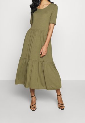 Frau trägt ein olivgrünes, gestuftes Midikleid mit kurzen Ärmeln und schwarze, knöchelhohe Sandalen mit Absatz, steht vor einfarbigem Hintergrund.