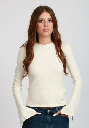Top lungo color beige chiaro con maniche a lunga, realizzato in un tessuto morbido. Presenta un corpo aderente e maniche svasate, con un collo a giro e una texture liscia.