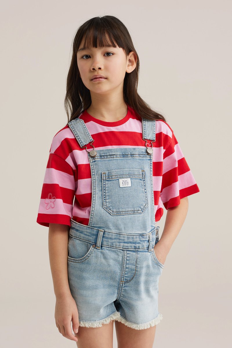Lány piros és rózsaszín csíkos rövid ujjú pólóban és világoskék farmer rövidnadrágos overállban, egyik kezét a zsebében tartva, egyszerű háttér előtt.
