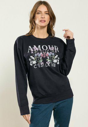 Donna che indossa una felpa nera con design floreale e testo "AMOUR C'EST LA VIE", abbinata a jeans blu, posando su uno sfondo neutro.