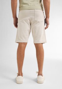 Homme debout de dos, portant un short beige, un t-shirt vert clair et des baskets gris clair, sur un fond uni clair.