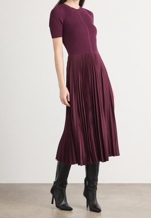 Nainen, joka pukeutunut luumunväriseen, lyhythihaiseen neulepaitaan ja siihen sopivaan laskostettuun midihameeseen sekä mustiin, korkokorkoisiin nahkasaappaisiin.