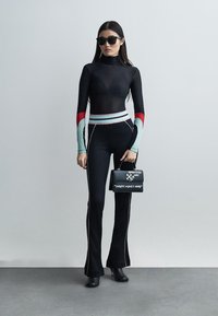 Γυναίκα με μαύρο εφαρμοστό ζιβάγκο με πολύχρωμα μανίκια, μαύρο καμπάνα παντελόνι, μαύρες μπότες, γυαλιά ηλίου και μαύρη τσάντα με λευκό κείμενο.