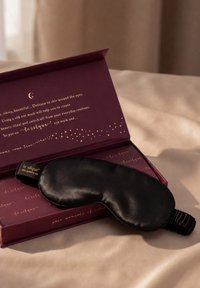 Zwart zijden slaapmasker rustend op een open donkerpaarse doos met gouden tekst en sterillustraties op een beige stoffen achtergrond.